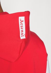 Red hoodie with a textured finish. Features a white label on the hood with the text "Arsenal" in a bold, black font.