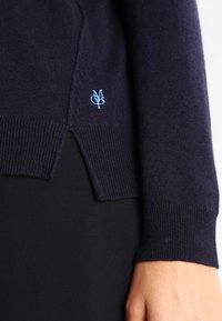 Navy blue sweater sleeve and side hem with light blue embroidered logo, worn over black pants, showing part of a person's hand and wrist.