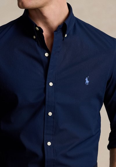 Mężczyzna ubrany w granatową koszulę zapinaną na guziki z zapiętym kołnierzykiem i jasnoniebieskim haftowanym logo na piersi.