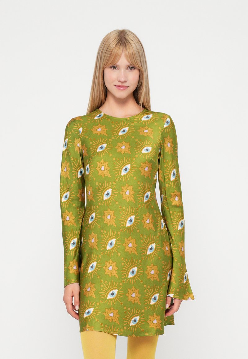 Grünes Tunika-Kleid mit langen Ärmeln, Augen- und Blumenmustern, ausgestellten Manschetten und weicher Textur. Getragen mit gelben Leggings.