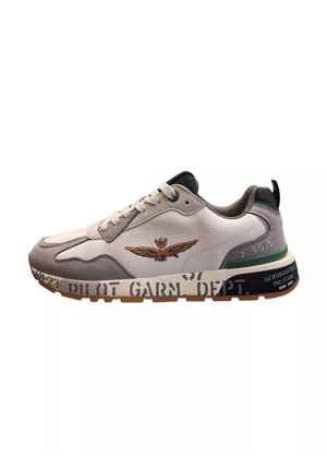 Sneaker bianco e grigio con materiali in suede e mesh, caratterizzato da un emblema alato dorato, accenti neri e dettagli testuali sulla suola.