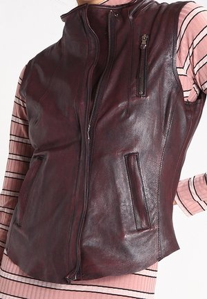 Mørkebrun lædervest med lynlås foran, lynlåslomme på brystet og to sidelommer, båret over en lyserød stribet langærmet skjorte.