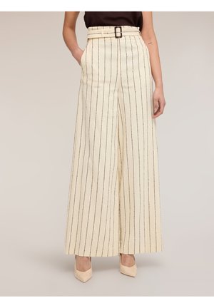 Kvinde iført højtaljede, viddebenede cremefarvede bukser med tynde sorte lodrette striber, sort bælte og beige spidse hæle.