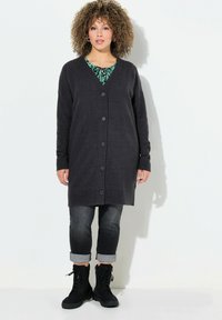 Dunkelgraue Strickjacke mit V-Ausschnitt, gerippten Bündchen und sechs Knöpfen. Getragen über einem grün gemusterten Oberteil, kombiniert mit hochgekrempelten Jeans und schwarzen Stiefeln.