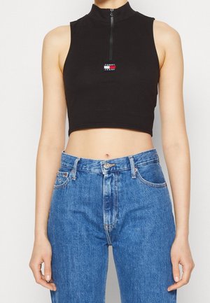 Mouwloze zwarte cropped top met rits aan de voorkant en logo, gecombineerd met een high-waisted blauwe jeans bij een persoon die voor een effen achtergrond staat.