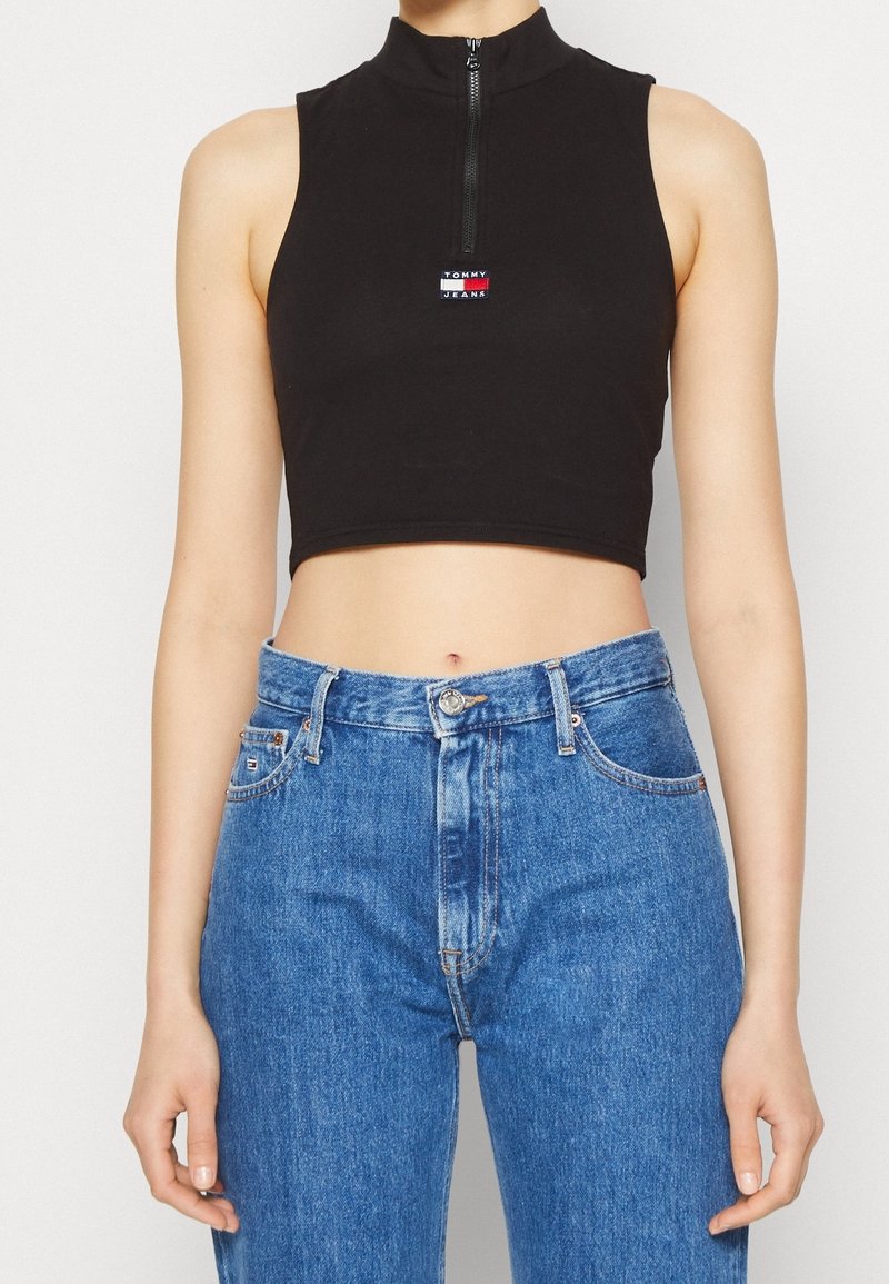 Haut court noir sans manches avec fermeture éclair sur le devant et logo, porté avec un jean bleu taille haute sur une personne debout devant un fond uni.