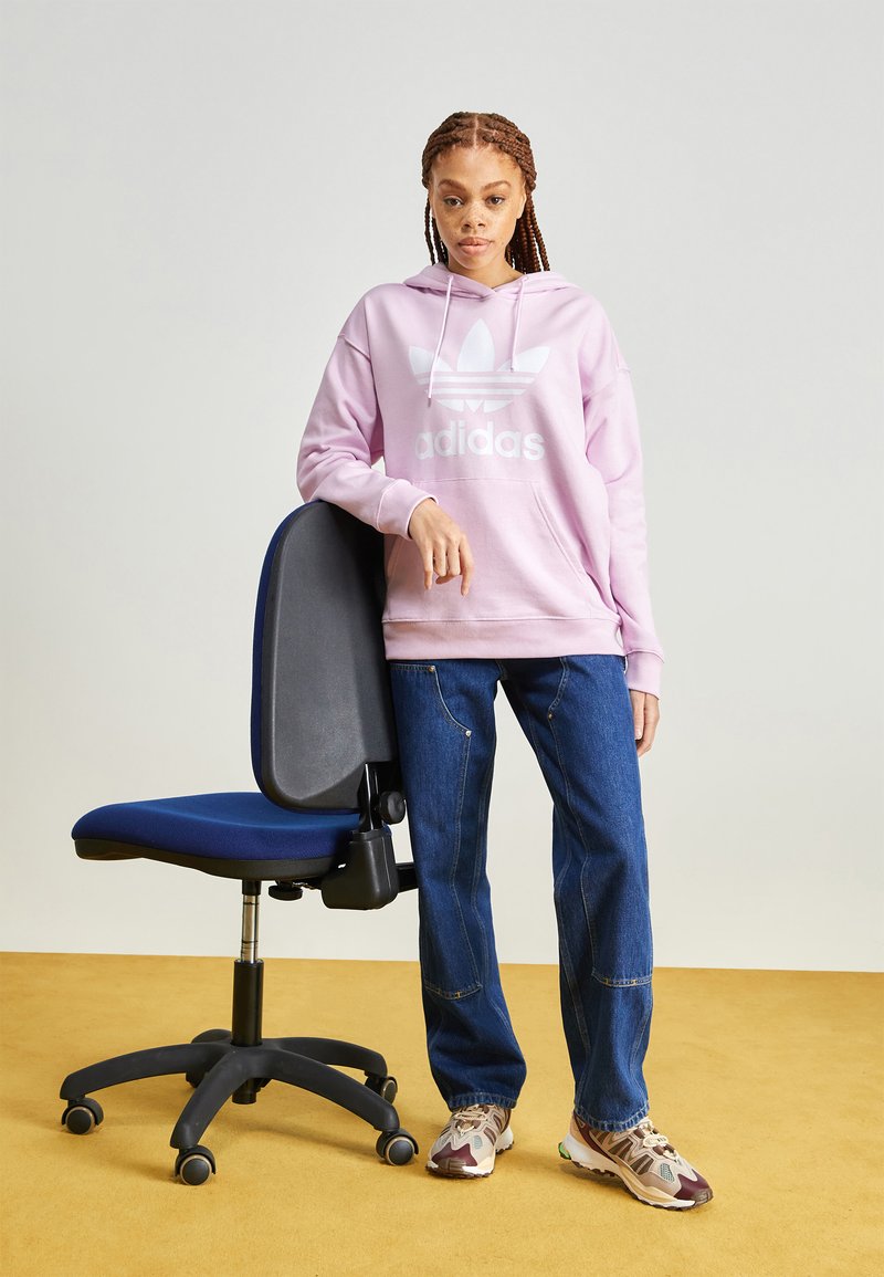 Rosa hettegenser med hvit logo, blå kontorstol, blå jeans og flerfargede joggesko. Glatt stoff og forskjellige teksturer er synlige.