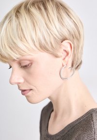 Boucles d'oreilles créoles en argent, texturées, présentant un design incurvé avec de petites pierres claires. Le modèle a les cheveux blonds courts, montré de profil.