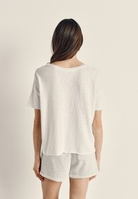 Žena s dlhými hnedými vlasmi stojí chrbtom, má na sebe voľné biele tričko a svetlošedé šortky, na jednoduchom pozadí.