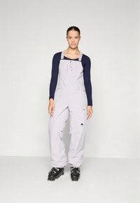 Macacão de esqui cinza claro com alças ajustáveis e bolsos, tecido texturizado. Combinado com uma blusa de manga longa azul-marinho e botas de esqui pretas.