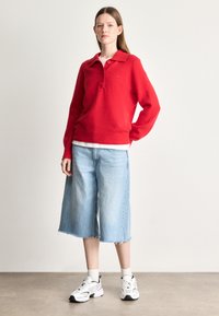 Červený vlnený sveter s golierom a dvoma gombíkmi, kombinovaný s svetlomodrými džínsovými šortkami s voľným strihom a bielymi teniskami so zelenými akcentmi.