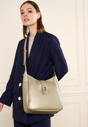 Jeune femme portant un blazer bleu marine et un top beige tenant un grand sac à épaule doré métallique avec détail de fermoir.