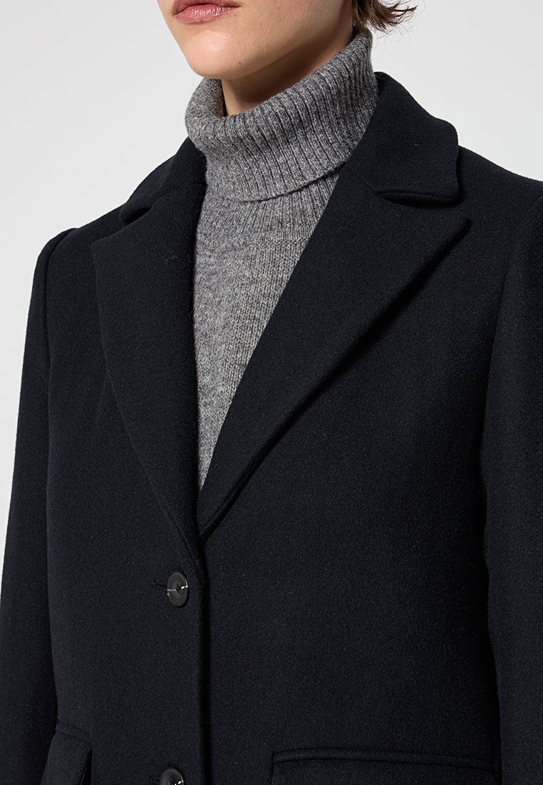 Manteau en laine mélangée bleu marine avec revers crantés, deux boutons et poches avant. Porté par-dessus un pull col roulé gris ajusté. Texture lisse.