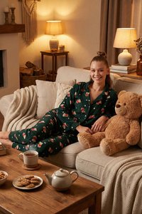 Frau in grünen, geblümten Schlafanzug entspannt auf einem beigen Sofa mit großem Teddybär, Holztisch mit Tee und Keksen, warme Lampen in einem gemütlichen Wohnzimmer.