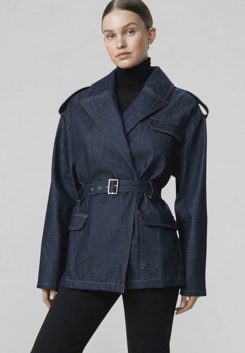 Žena nosi tmavú rifľovú bundu s opaskom, veľkým golierom a vreckami s chlopňami, v kombinácii s čiernymi nohavicami a čiernym rolákom.