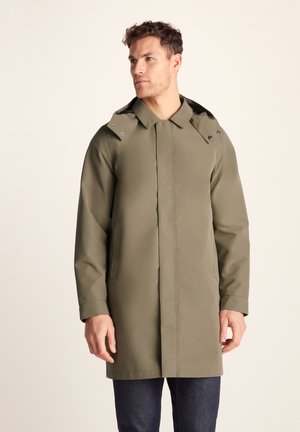 Man wearing a long olive green hooded coat with concealed buttons, standing against a plain light background.