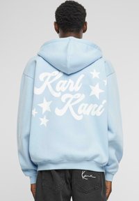 Helle blaue Kapuzenjacke mit weißem "Karl Kani"-Text und Sternen-Grafiken auf der Rückseite; ausgestattet mit einer Kordelzugkapuze und gerippten Bündchen.