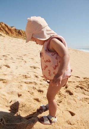 Vaaleanpunainen uimaliivi merihevosen kuvioinnilla, raidallinen aurinkohattu ja vaaleat sandaalit. Lapsi seisoo hiekkarannalla katsoen alaspäin.