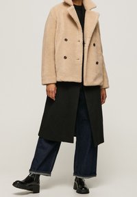 Osoba nosząca beżowy dwurzędowy płaszcz teddy na długim czarnym płaszczu, ciemne spodnie z szerokimi nogawkami oraz czarne masywne botki.