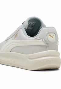Zapatilla en gris y blanco con parte superior de cuero suave y acolchado blando. Presenta la marca PUMA y una suela de goma texturizada.