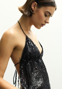 Sort palietkjole med halterneck og dyb V-udskæring, der har tynde stropper og en flydende silhuet. Teksturen er skinnende med en glitrende effekt.