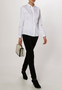Donna che cammina indossando una camicia bianca aderente con bottoni, pantaloni neri, scarpe con tacco nere e portando una borsa beige con una maniglia a catena dorata.