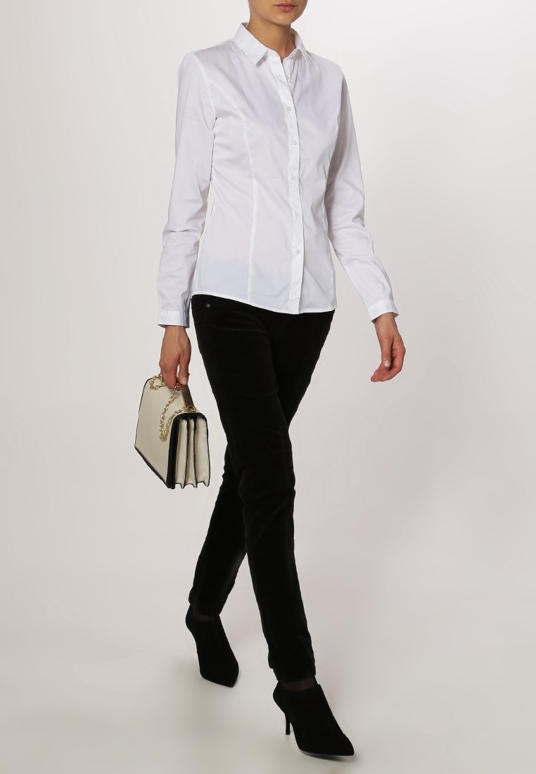 Donna che cammina indossando una camicia bianca aderente con bottoni, pantaloni neri, scarpe con tacco nere e portando una borsa beige con una maniglia a catena dorata.