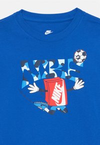 Kék pamut póló, melyen egy játékos grafika látható egy karakterről, aki egy piros dobozt és focilabdát tart, a "NIKE" felirat stilizált, különböző színekben.