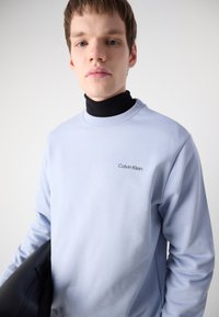 Fénykék pulóver, kerek nyakkal, fekete logóval a bal felső sarokban, fekete garbó felett viselve, fekete kiegészítővel rögzítve.
