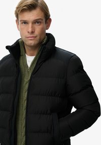 Chaqueta acolchada negra con cuello alto y bolsillos laterales. Usada sobre un suéter verde de punto cableado, con un acabado liso y mate.
