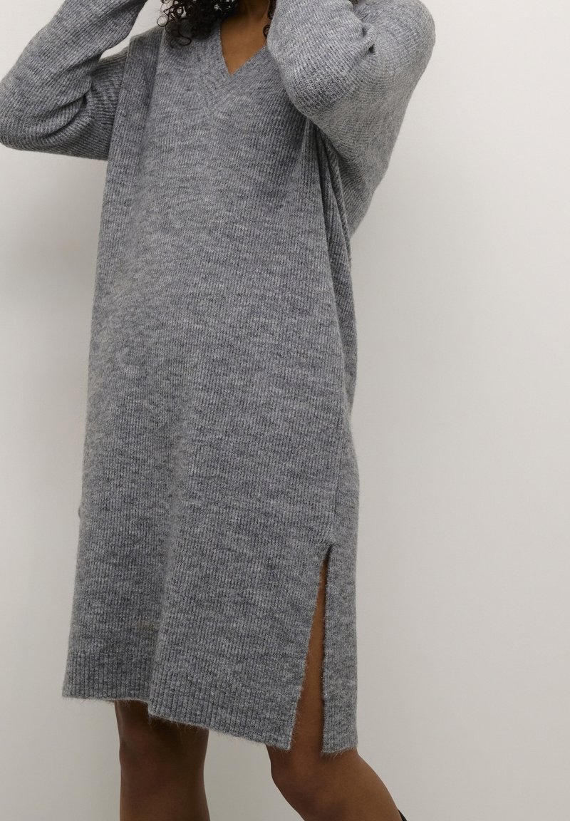 Femme portant une robe-pull en maille grise avec une fente latérale, debout devant un fond uni de couleur claire.