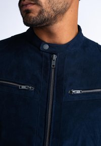 Giacca in suede blu navy con colletto alto, chiusura frontale con zip e due tasche sul petto con zip. Presenta un tessuto testurizzato e dettagli di cucitura.