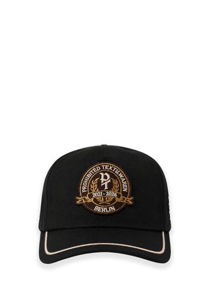 Jeune homme portant une casquette noire avec un emblème, un sweat à capuche noir et une veste en velours côtelé marron, regardant pensivement sur le côté sur un fond uni.