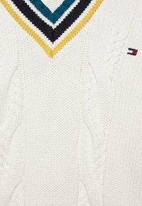 Biały sweter w splot warkoczowy z dekoltem w serek, z żółtymi, czarnymi i turkusowymi paskami oraz małym czerwono-białym-czarnym logo na rękawie.