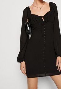 Svart miniklänning med en texturerad tyg, puffade långa ärmar, fyrkantig halsringning och framknappdetaljer. Accenterad med en liten handväska.