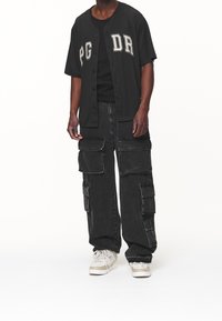 Persona che indossa una maglietta da baseball nera con bottoni e la scritta "PG DR", pantaloni cargo neri con grandi tasche e sneaker bianche, in piedi dritto.