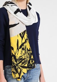 Écharpe légère et tissée en blanc, jaune et noir avec un imprimé botanique ; se drape en boucle autour du cou, avec une texture douce.