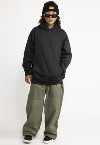 Zwarte hoodie met een voorzak en een tonale logo, gecombineerd met losse olijfgroene broeken. Zwarte sneakers en een zwarte muts met een label.
