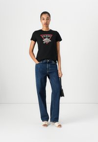 Svart bomull T-skjorte med rød trekantet grafikk og blomsterdesign, kombinert med løse mørke vaskede jeans og kremfargede sandaler med åpen tå.