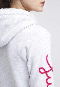 Sudadera gris claro con una textura suave, con un gran logotipo en cursiva rosa en el brazo izquierdo y una capucha estándar.