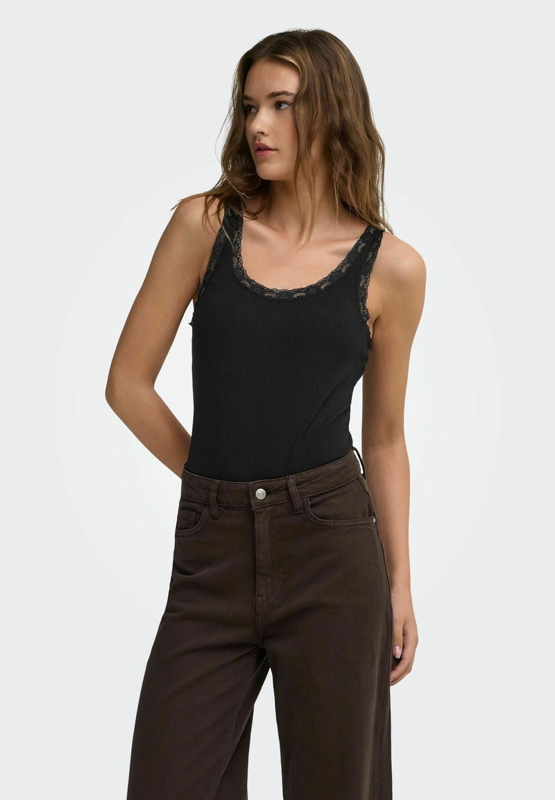 Femme aux cheveux bruns mi-longs, vêtue d'un haut sans manches noir orné de dentelle et d'un pantalon marron foncé taille haute, regardant sur le côté.