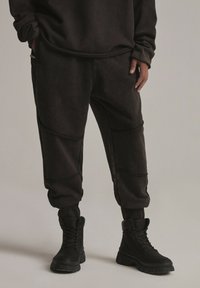 Ciemnobrązowe dresy o luźnym kroju, z zaokrąglonymi szwami i elastycznymi mankietami. Połączone z czarnymi sznurowanymi butami dla spójnego wyglądu.