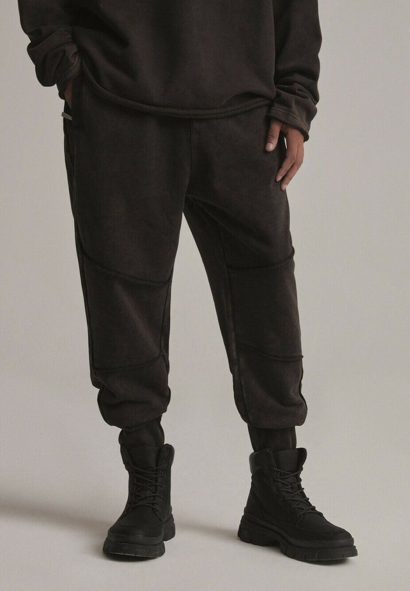 Ciemnobrązowe dresy o luźnym kroju, z zaokrąglonymi szwami i elastycznymi mankietami. Połączone z czarnymi sznurowanymi butami dla spójnego wyglądu.