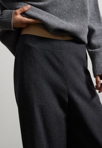 Maglione di lana grigio leggermente sollevato per rivelare pantaloni di lana grigio scuro a vita alta, con una texture liscia e un design minimal.