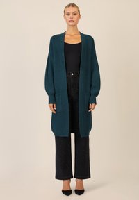 Cardigan lavorato a maglia teal con spalle cadenti, apertura frontale e due tasche laterali, indossato sopra una maglietta nera e pantaloni in denim scuro.