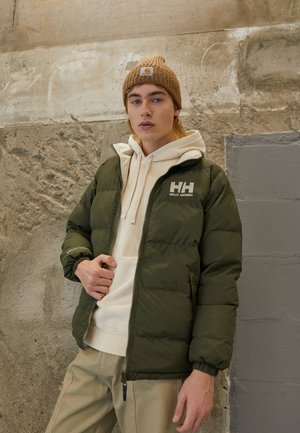 Zelená prešívaná bunda s bielym logom, skombinovaná s smotanovou mikinou a svetlými khaki nohavicami. Model má na sebe hnedý pletený čiapku.