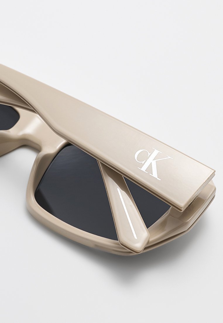 Beżowe okulary przeciwsłoneczne o kanciastym kształcie, z czarnymi szkłami i metalicznym logo "cK" na zauszniku. Posiadają błyszczące wykończenie oraz unikalny kształt.