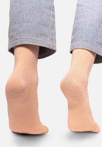 Beżowe skarpetki ankle o gładkiej fakturze i wzmocnionym palcem. Jedna skarpetka ma żółty akcent na palcu. Noszone z podwiniętymi jasnoniebieskimi dżinsami.
