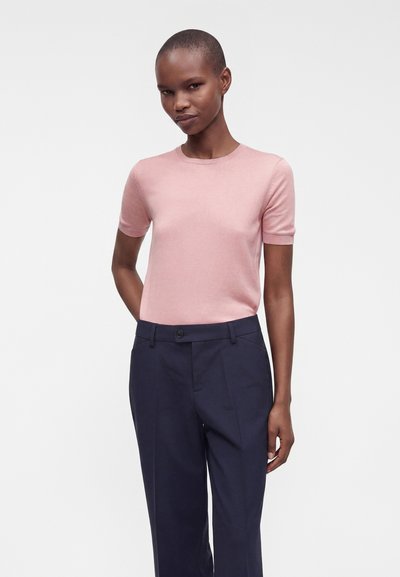 Femme portant un pull rose clair à manches courtes et un pantalon bleu marine foncé sur mesure, debout devant un fond blanc uni.