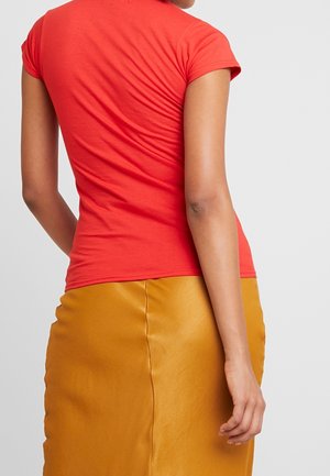 Nainen pukeutuneena istuvaan punaiseen lyhythihaiseen paitaan ja sinapinkeltaiseen nahkahameeseen, nähtynä takaa yksinkertaista vaaleaa taustaa vasten.